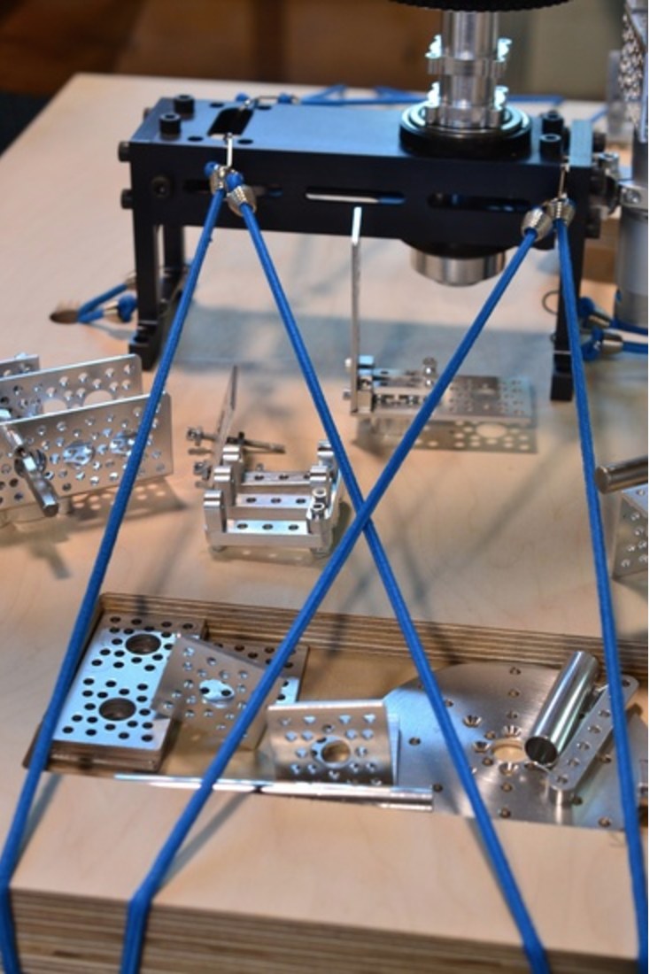Photograph of machined aluminium art-machine components laid out on a birch surface beneath a black frame, with blue elastic cords crossing in tension in the foreground